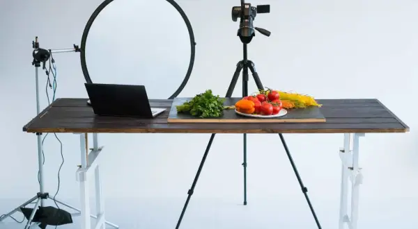 A tabletop photography setup with a camera on a tripod, a laptop, and fresh ingredients like tomatoes and pasta on a wooden table.