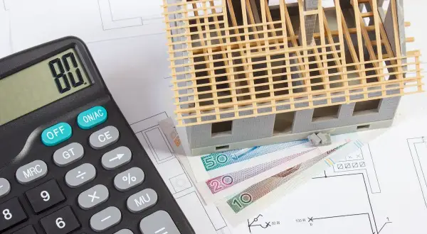 Kalkulator, makieta domu wielorodzinnego i polskie banknoty na planach budowlanych. Zastanawiasz się, ile kosztuje budowa domu wielorodzinnego?