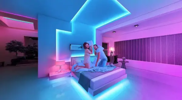 A couple has a pillow fight in a bedroom bathed in vibrant RGB light ideas, with blue and pink hues illuminating the modern space.
