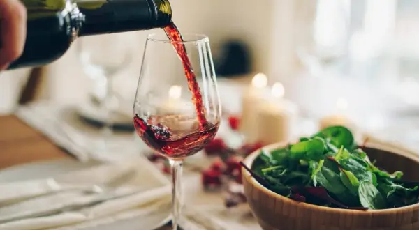 Versando vino rosso in un bicchiere, con un'insalata fresca e candele sullo sfondo. Un'atmosfera perfetta per gustare un buon greco di tufo vino.