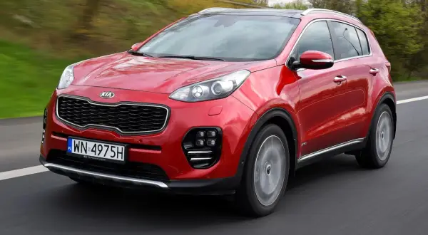 Czerwony Kia Sportage IV pędzi po drodze, otoczony zielenią.