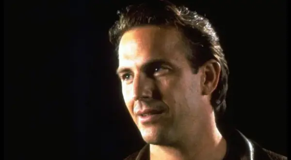 Kevin Costner jung, mit nachdenklichem Blick und brauner Lederjacke vor dunklem Hintergrund.