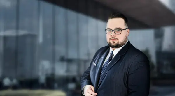 Mężczyzna w garniturze i okularach na tle nowoczesnego budynku.
