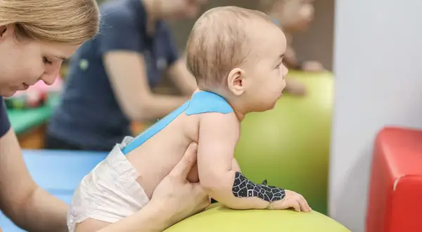 Dziecko na piłce rehabilitacyjnej, z taśmami kinesiotaping. Terapeuta wspiera malucha podczas ćwiczeń.