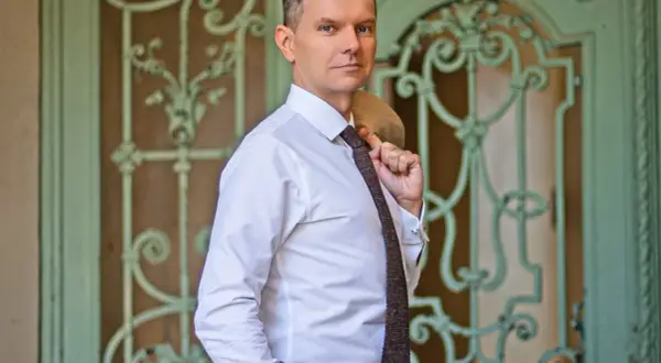 Tomasz Roy Szabelewski w białej koszuli i krawacie, z marynarką na ramieniu, przed ozdobnymi drzwiami.