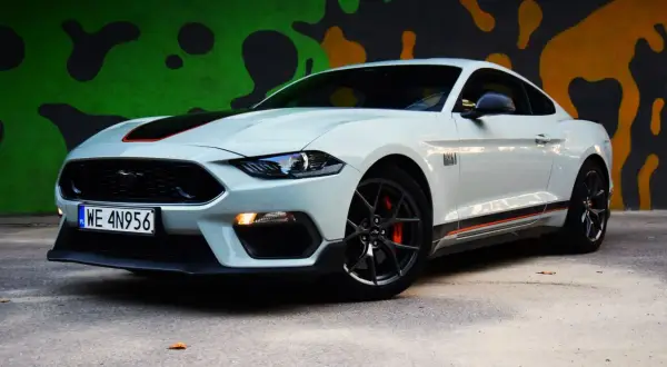 Biały Ford Mustang Mach 1 z czarnymi pasami i pomarańczowymi akcentami na tle kolorowego muralu.