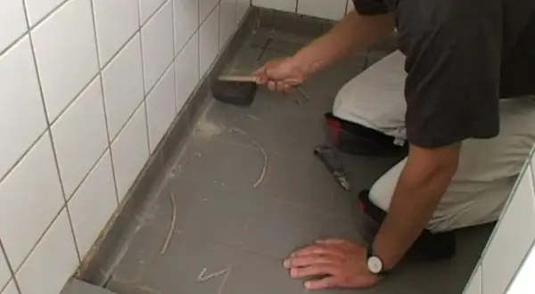 Man knäböjer och använder en borste för att ta bort silikonfog runt en dusch.