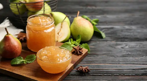 Confiture de poires dans un bocal et un bol, avec des poires fraîches et des feuilles de menthe sur une planche en bois.