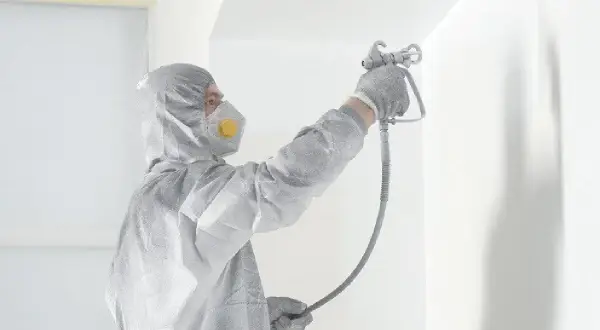 Osoba w kombinezonie i masce ochronnej wykonuje malowanie pistoletem białej ściany.