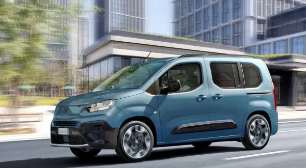 Un Fiat Doblò 9 posti blu metallizzato sfreccia in città, con grattacieli moderni sullo sfondo.