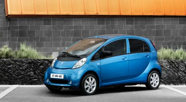 En blå Peugeot iOn står parkerad framför en grå tegelvägg med en orange takkant.