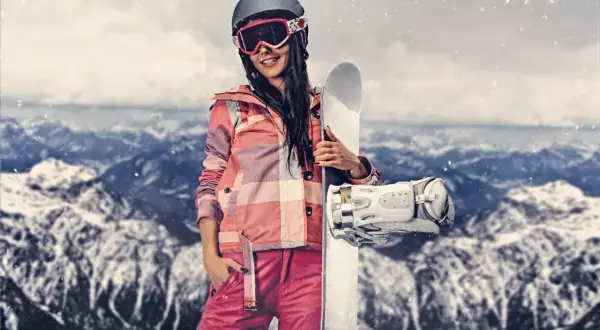 Kobieta w stroju narciarskim trzymająca snowboard na tle gór.