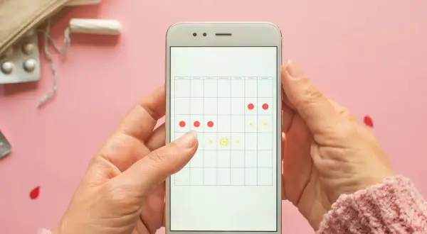 Aplikacja na telefonie pokazuje kalendarz cyklu. Czy można iść na wizytę do ginekologa podczas okresu? Na zdjęciu widać też tampony i tabletki.