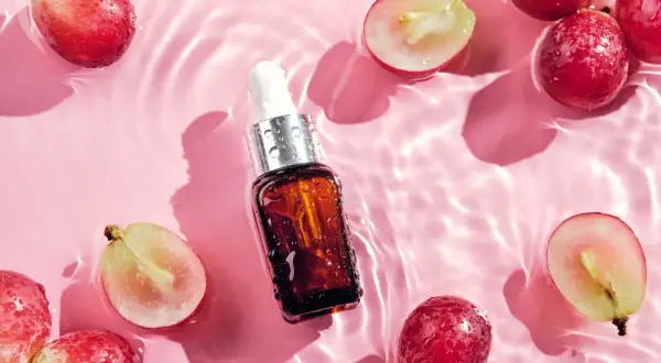 Odkryj naturalne olej z pestek winogron właściwości. Serum w buteleczce z pipetą otoczone świeżymi winogronami na różowym tle z falami wody.