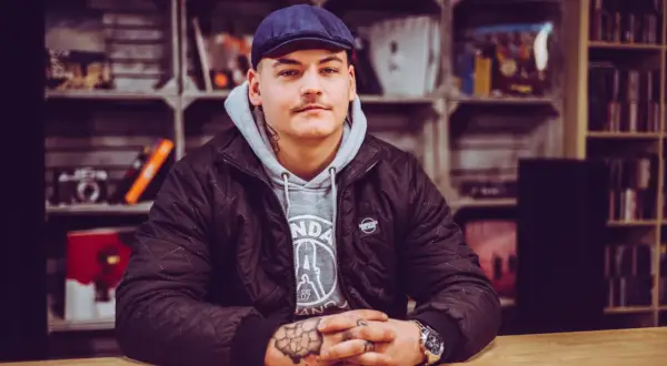 Młody mężczyzna w czapce i kurtce, z tatuażami na rękach, siedzi przy drewnianym stole. Wygląda jakby przeżywał swój **frosti wiek**, pełen pasji i determinacji.