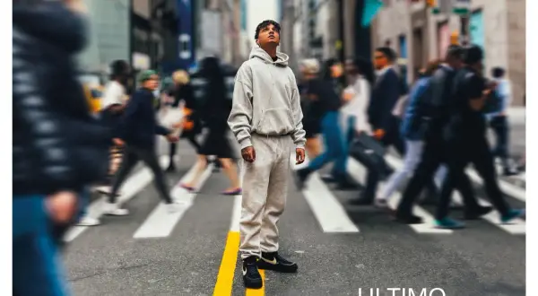 A young man in a grey tracksuit stands on a crosswalk in a busy city street, looking up.