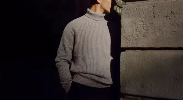 Uomo con un elegante outfit con dolcevita grigio, in piedi vicino a un muro di pietra.