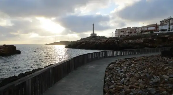 Camino de piedra y madera bordeando el mar, con un faro al fondo. La carretera subida al faro invita a pasear al atardecer.