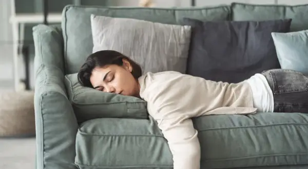 Eine Frau liegt erschöpft auf einem Sofa, ihre Augen sind geschlossen. Sie leidet unter Gliederschmerzen ohne Fieber und ruht sich aus.