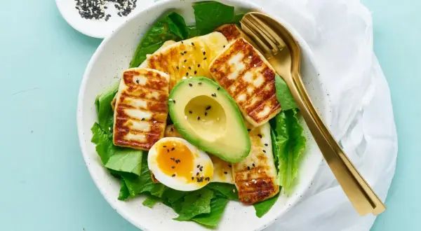 Idealne keto śniadanie: grillowany ser halloumi, awokado, jajko na miękko i świeża sałata, posypane czarnym sezamem.