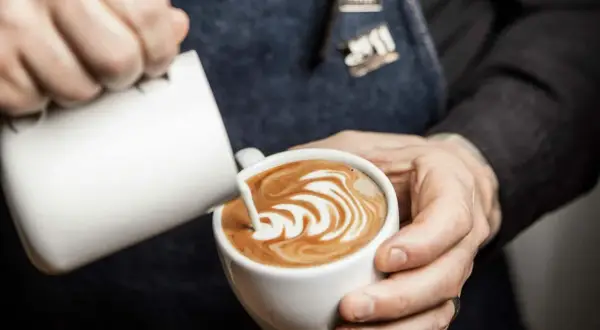 Barista skapar vacker latte art i en vit kopp. Mjölkskummet bildar ett mönster av virvlar.