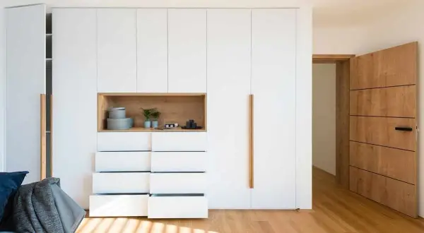 Armarios ocultos en pared blancos con nicho de madera y cajones abiertos. Puerta de madera a la derecha.