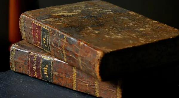 Dwa stare, skórzane tomy, jeden na drugim. Na grzbietach widoczne złote zdobienia i etykiety z łacińskimi napisami, sugerujące treść jak psalm 98.