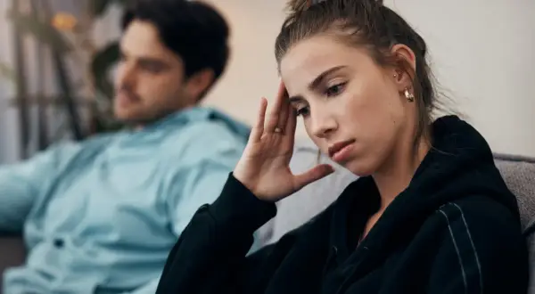 Zmartwiona kobieta z ręką na czole, podczas gdy jej mąż wmawia jej chorobę psychiczną.