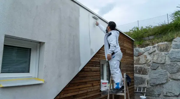 Mann streicht Fassade ohne Reinigung mit Roller. Ein Eimer Farbe steht bereit.