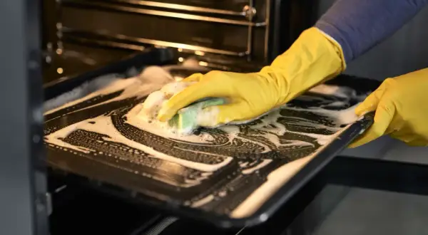 Main tenant avec des gants jaunes, nettoie une plaque de cuisson savonneuse pour enlever odeur four.