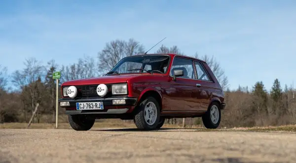 Röd Peugeot 104 med extraljus parkerad på en grusväg, med träd och blå himmel i bakgrunden.