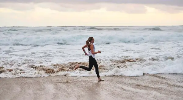 Frau joggt barfuß am Strand entlang, während die Wellen des Meeres an den Sand spülen. Sie genießt ihr tägliches Joggen.