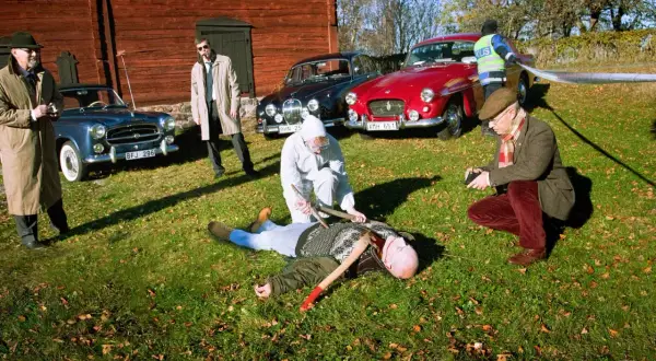 En brottsplats med en död man på marken, omgiven av gamla bilar, inklusive en British Leyland. Polis och utredare arbetar.