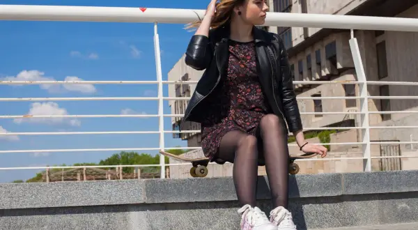 Dziewczyna w skórzanej kurtce i kwiecistej sukience, siedząca na deskorolce, prezentuje modne stylizacje z butami sportowymi.