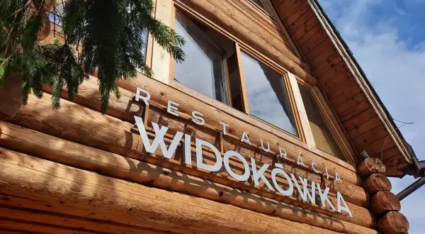 Drewniana restauracja Widokówka z białym napisem. Gałązka świerku zwisa z lewej strony.