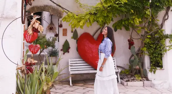 Una donna sorride sotto un albero rigoglioso, con un grande cuore rosso e decorazioni che ricordano il pomodoro ruffano.