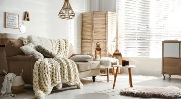 Przytulny salon w stylu hygge wnętrza. Beżowa kanapa z grubym, plecionym kocem, wiklinowa lampa, parawan i świece tworzą ciepłą atmosferę.