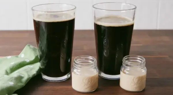 Zwei Gläser dunkles Bier und zwei kleine Gläser mit cremiger Flüssigkeit auf einem Holztisch.