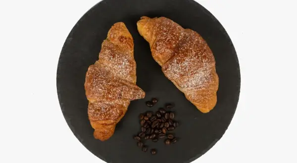 Dos cruasanes dorados espolvoreados con azúcar glas, junto a granos de café. Una delicia para aprender como hacer cruasanes.