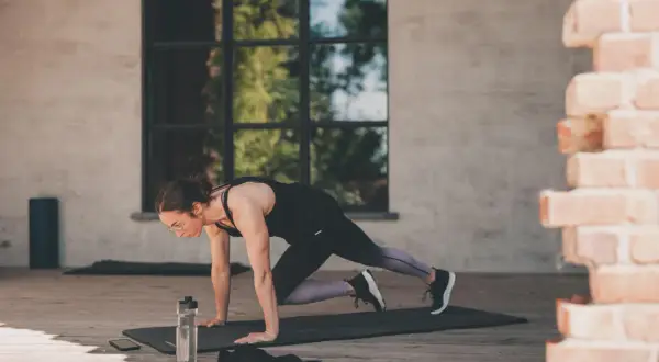 Femme en position de planche, genou vers poitrine, effectuant un exercice polyarticulaire. Bouteille d'eau et téléphone à proximité.