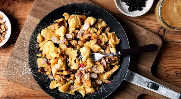 Kaiserschmarrn-Rezepte: Ein goldbrauner Kaiserschmarrn mit Mandelsplittern, Rosinen und Apfelstücken in einer Pfanne, bestäubt mit Puderzucker.