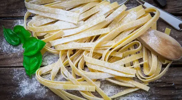 Świeży, domowy makaron bez glutenu, posypany mąką, z listkami bazylii i drewnianą łyżką na drewnianym blacie.