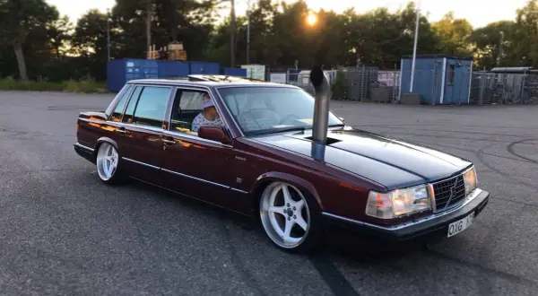 En mörkröd Volvo 760 D24 med en vertikal avgasrör som sticker upp från motorhuven. Bilen har vita fälgar och är parkerad på en asfalterad yta.