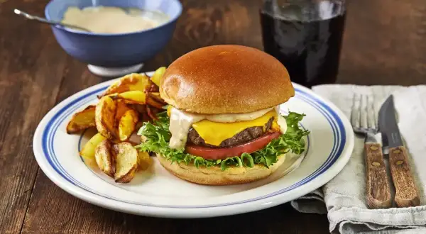 En saftig hamburgare med ost, sallad och tomat, serverad med klyftpotatis och en skål med krämig hamburgerdressing.