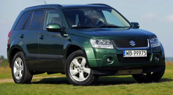 Zielony Suzuki Grand Vitara na tle łąki i błękitnego nieba. Kierowca za kierownicą.