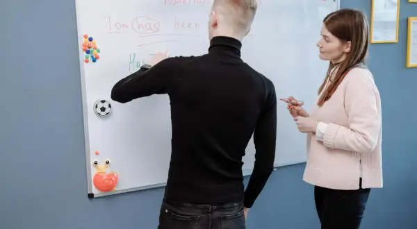 Nauczycielka i uczeń omawiają na tablicy zaimki, pisząc "Tom has been".