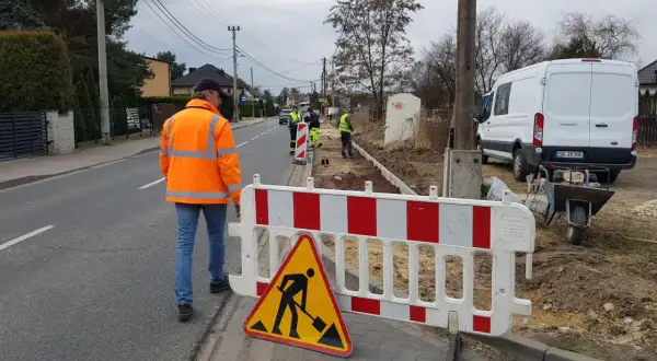 Robotnicy w kamizelkach odblaskowych pracują na budowie przy drodze, ogrodzonej barierkami.