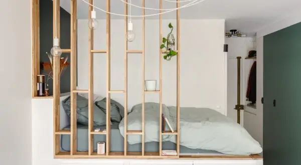 Un studio avec un coin nuit séparé par une étagère en bois. Le lit est fait de draps verts pâles.