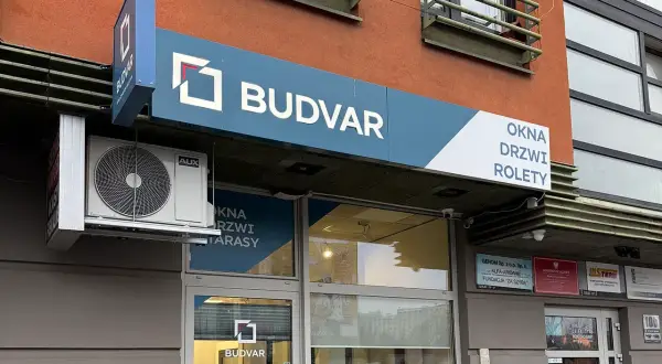 Logo firmy BUDVAR z napisem "OKNA DRZWI ROLETY" na tle pomarańczowej ściany.