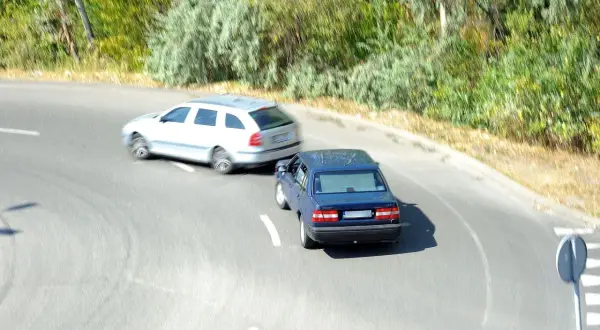 Niebieski samochód wyprzedza białe kombi na zakręcie, ryzykując wymuszenie pierwszeństwa i mandat.